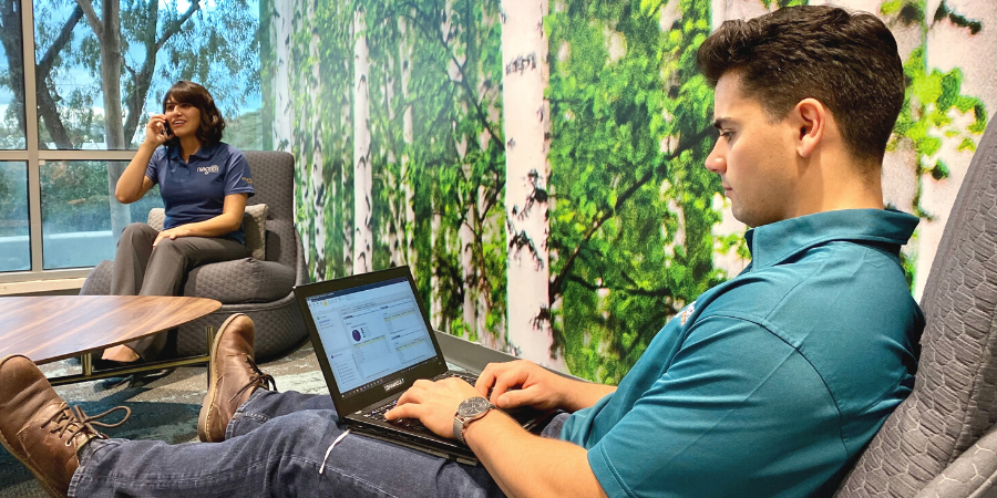 Man sitting in a comfortable chair while typing on his computer; woman in the background on her phone.