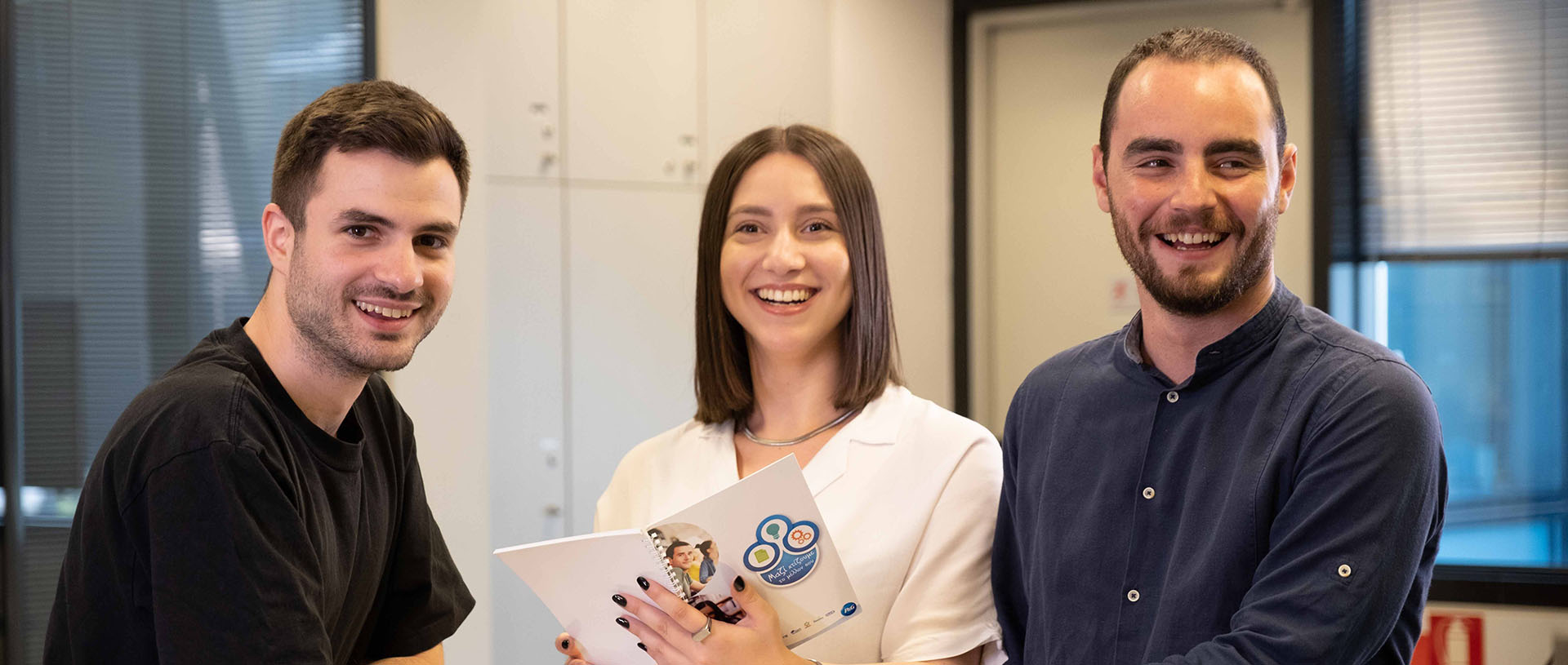 People smiling, holding a pamphlet, in an office setting.