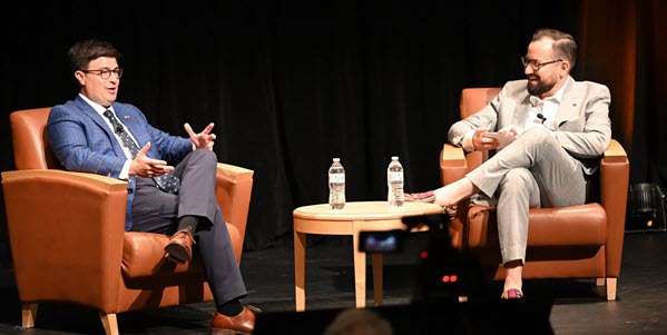 Two men sitting in arm chairs talking with each other on stage