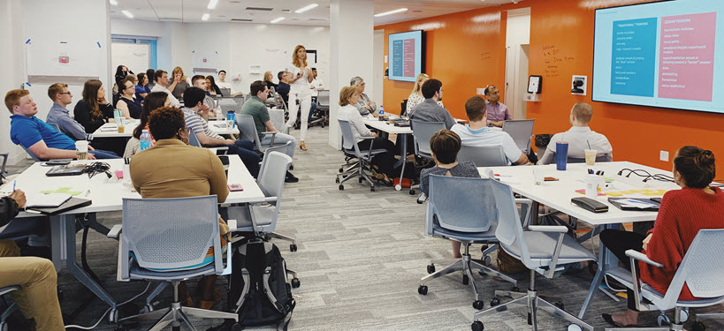 A picture of a conference room with groups of people at tables reviewing information on a tv screen on the wall