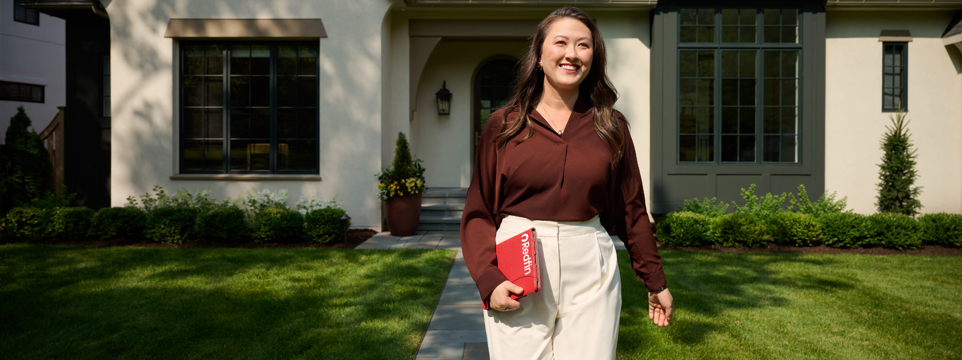 A smiling Redfin real estate agent walks confidently along a front path toward the camera, holding a red Redfin-branded folder. She is in front of a well-maintained, modern home with large windows and neatly trimmed landscaping on a sunny day.