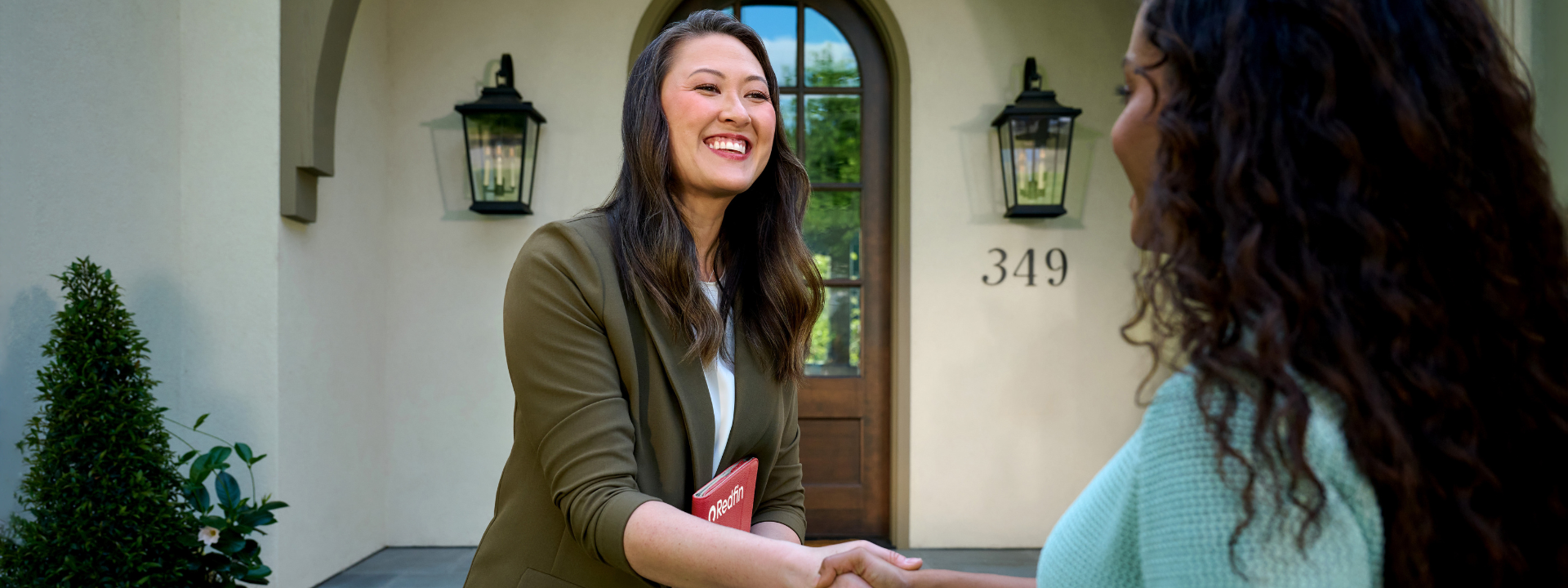 A cheerful Redfin real estate agent greets a client with a handshake in front of a house numbered 349. The agent, holding a red Redfin-branded folder, smiles warmly during the outdoor meeting. The home’s entrance features large lantern-style lights and arched architectural details.