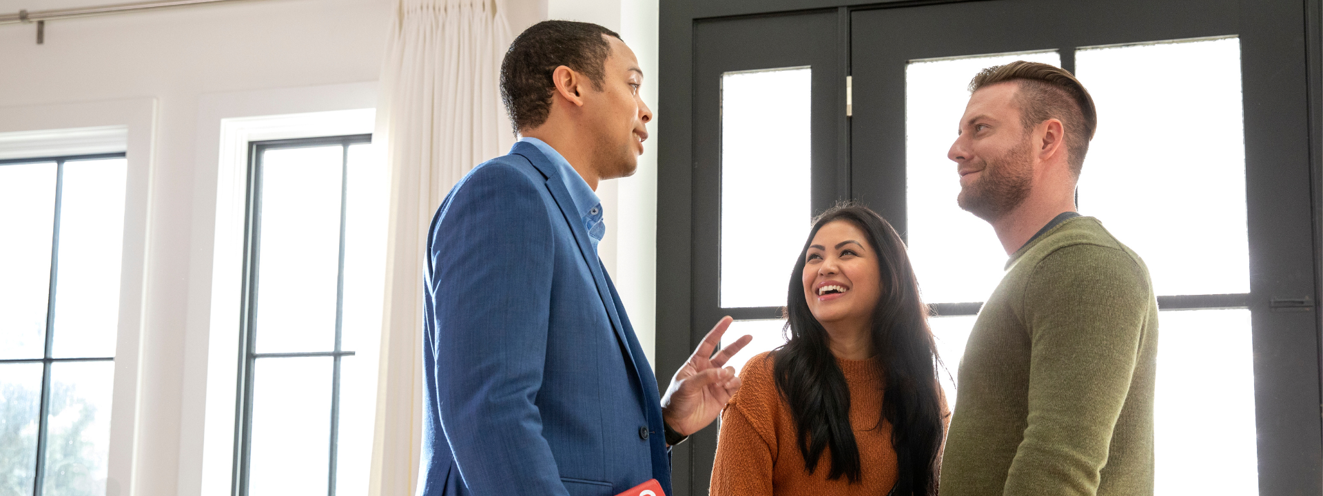 A Redfin agent in a blue suit talks with a smiling couple inside a brightly lit home. The couple listens attentively and appears engaged, standing near large windows and a modern black front door. The agent holds a red Redfin folder at his side.