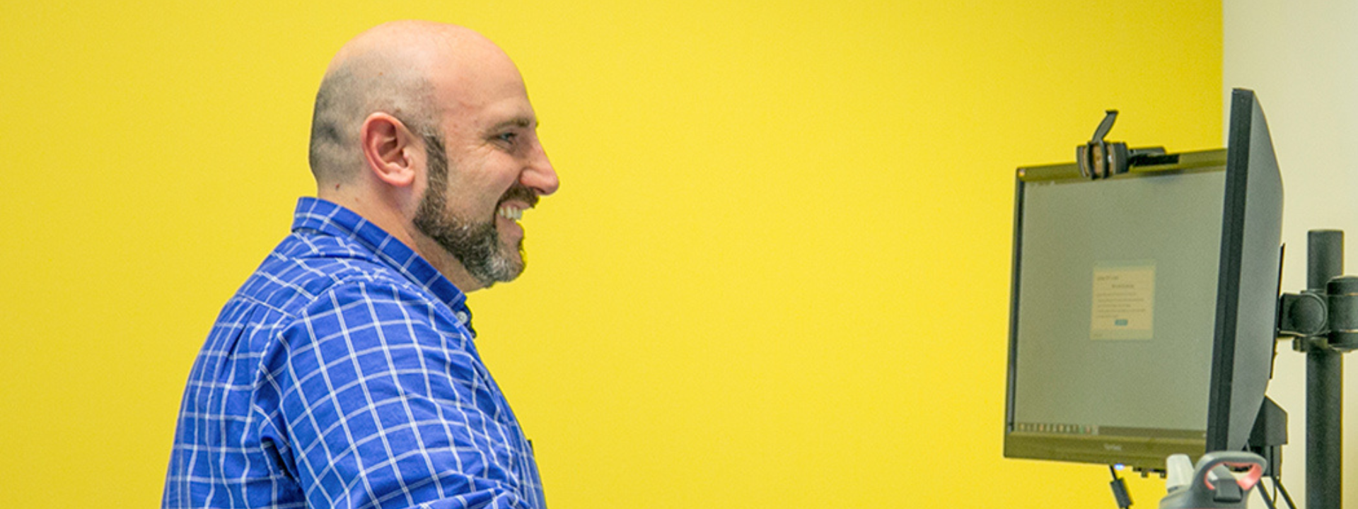 A smiling man in a blue checkered shirt works at a standing desk in front of a bright yellow wall. He types on a keyboard while looking at a monitor, with a water bottle featuring a Chicago Cubs sleeve placed on the desk beside him.