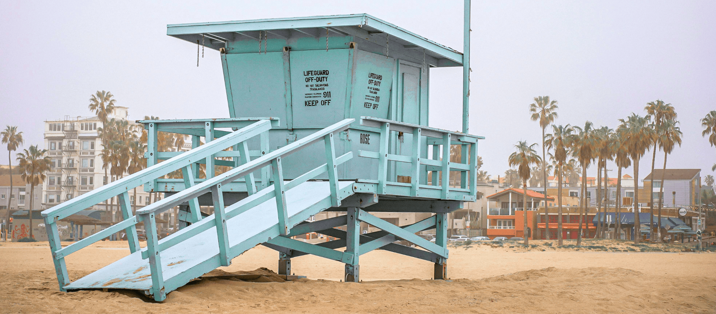 Pale blue lifeguard tower on a foggy morning at Venice Beach with palm trees and buildings in the background.