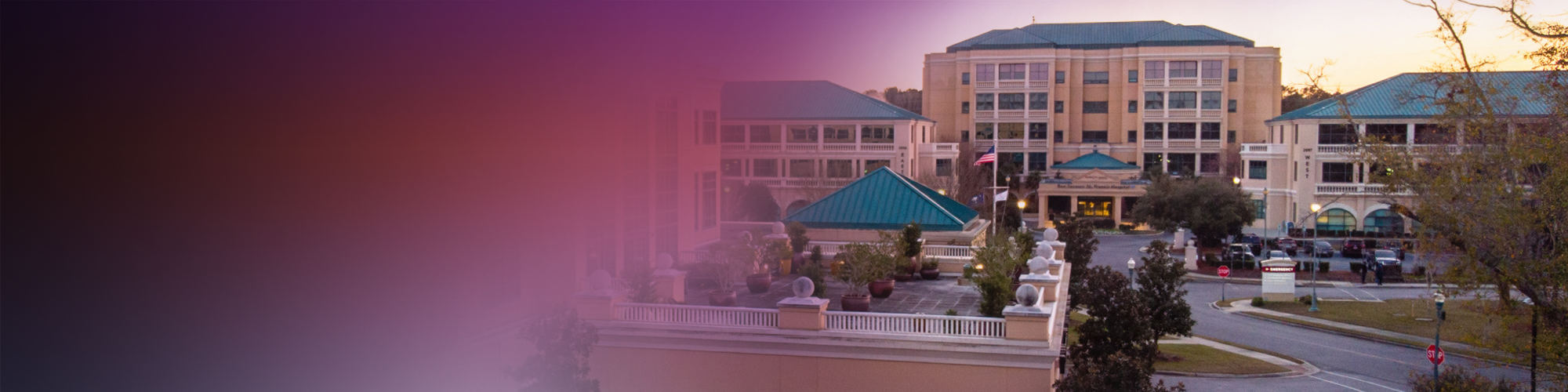 Exterior view of a large hospital or medical center at dusk, featuring multiple buildings with green rooftops, a landscaped terrace, and an entrance with an American flag.