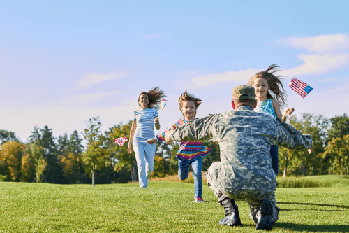 a veteran in camo uniform squatting down waiting for three children running up to hug him