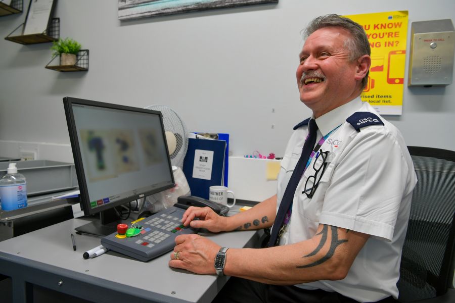 A man in a white shirt and Serco uniform sat at a desk
