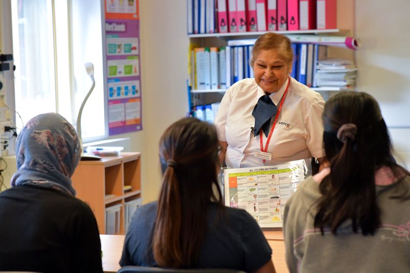 A woman in a white shirt and blue tie speaking to three people and showing them a guide.