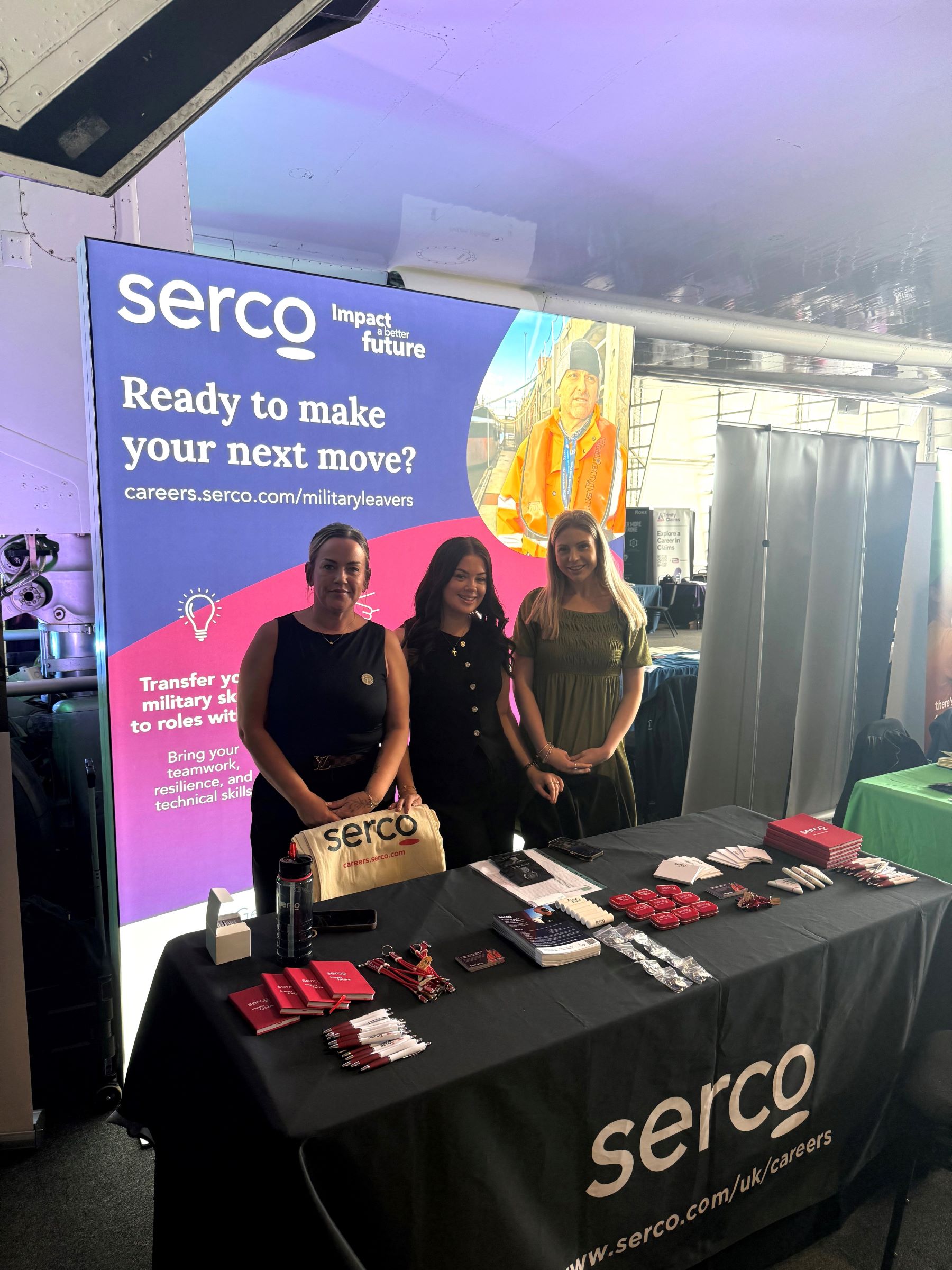Three colleagues at a recruitment event stand with Serco branding and merchandise.