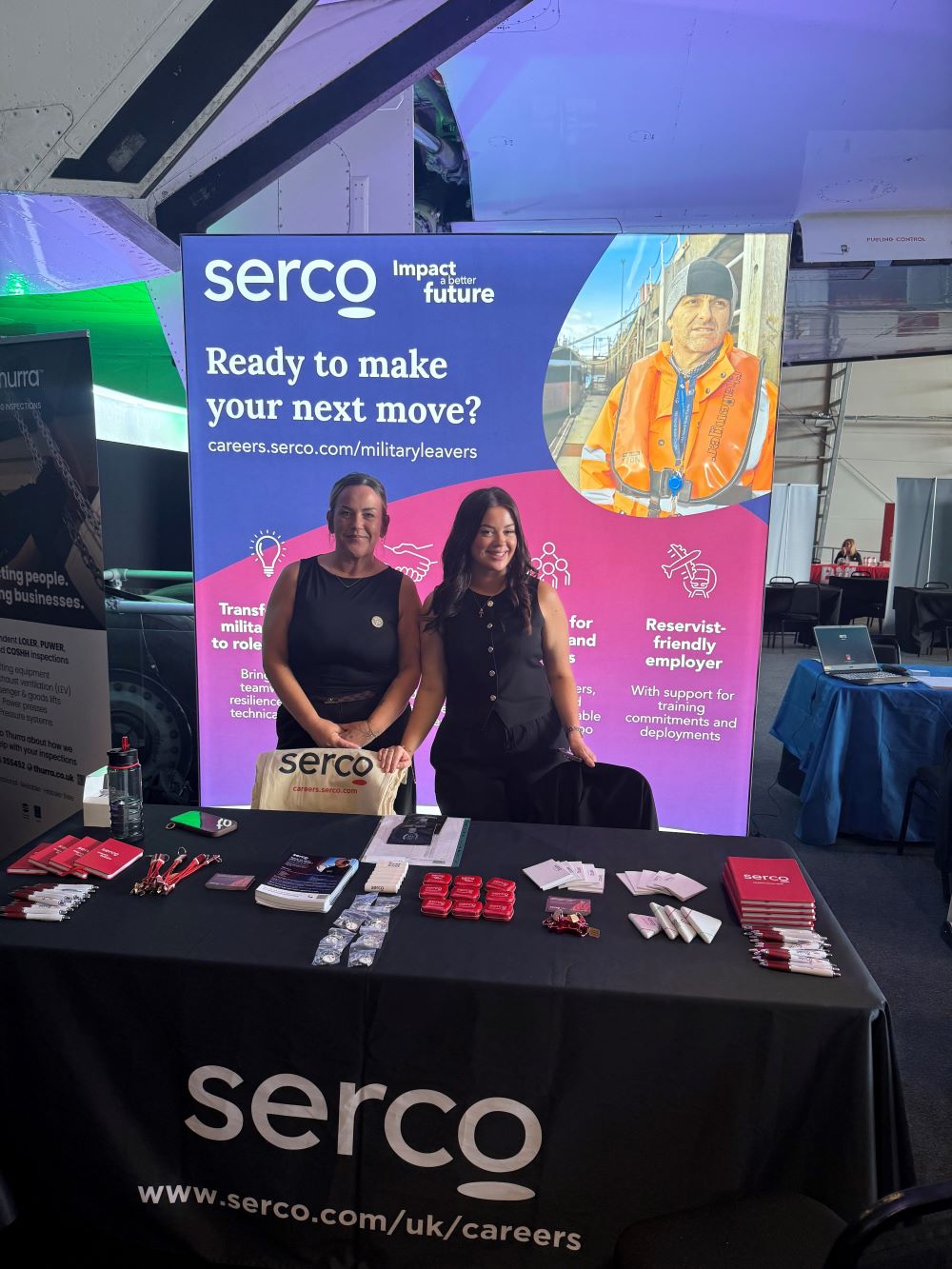 Two women at a careers event stand that says Serco and had merchandise on it