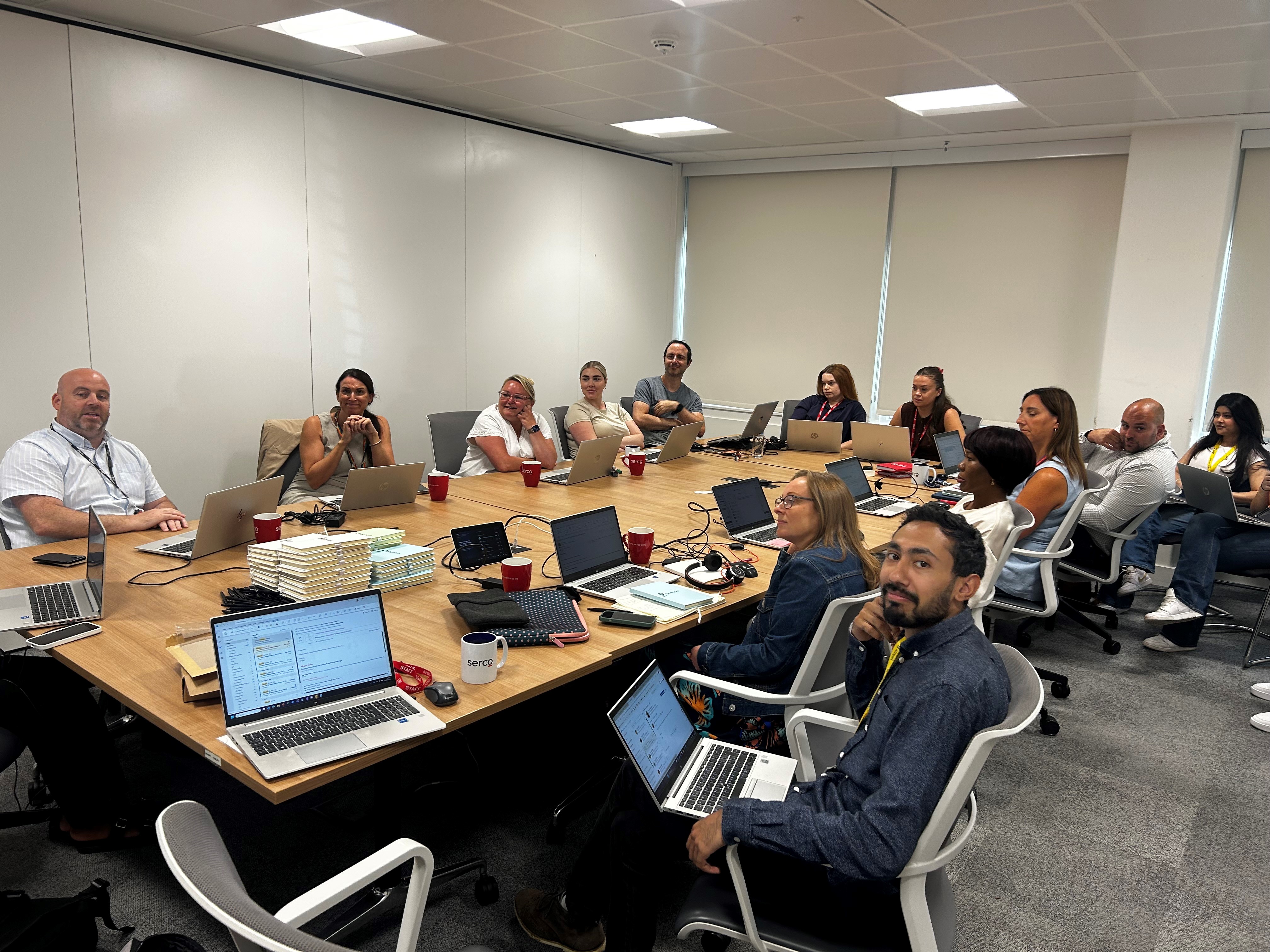 A group of people sat around a large table with laptops, smiling at the camera