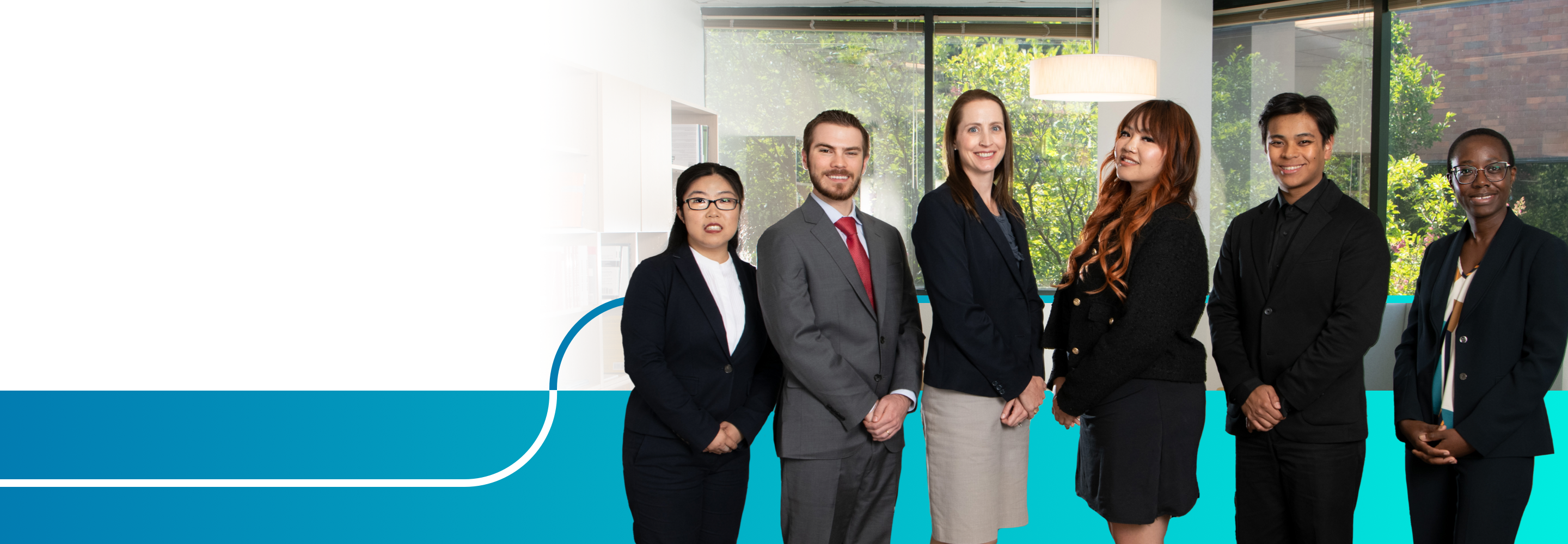 A group of six professionally dressed people standing indoors. They are in a modern office setting, with a bookshelf on the left and large windows showing green foliage in the background. The individuals are smiling and standing in a line, facing the camera.