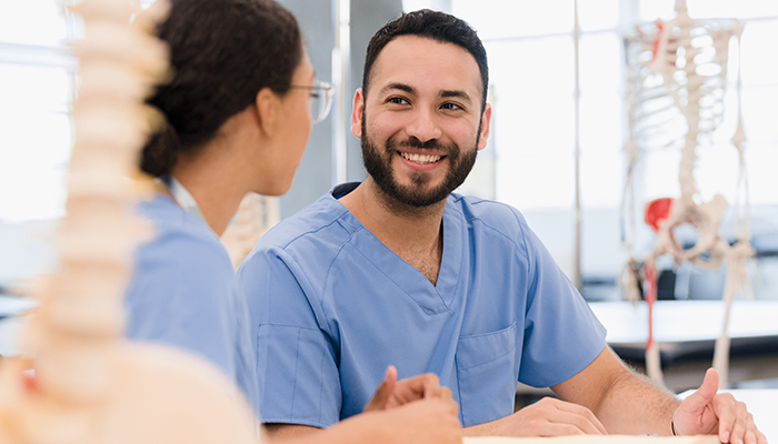 Two healthcare professionals in blue scrubs are having a conversation in a medical setting with anatomical models in the background. One individual, with glasses and hair tied back, faces a bearded colleague who is smiling.