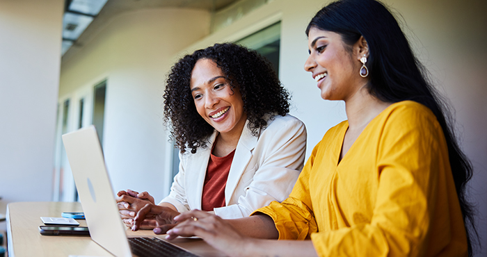 Two women are sitting at a table and smiling while looking at a laptop.