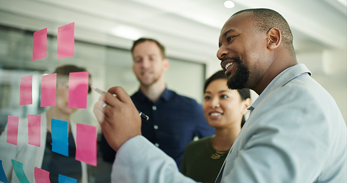 A group of four diverse professionals collaborates in an office. One man is writing on a glass wall covered with pink and blue sticky notes, while three colleagues stand behind him, engaged and smiling. The atmosphere appears productive and positive.