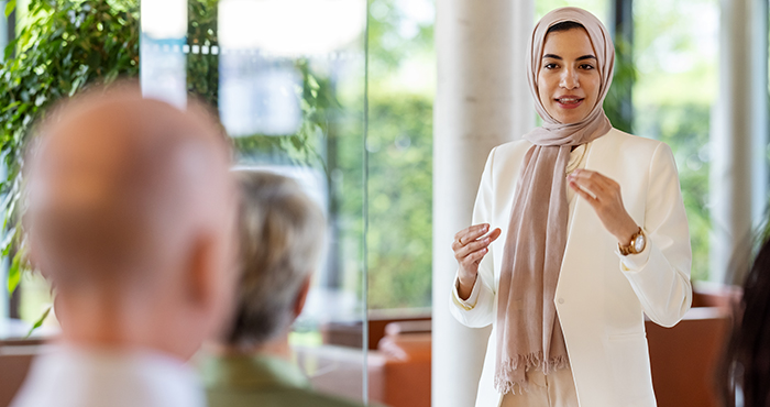 A woman wearing a hijab and business attire stands and speaks confidently in front of an audience in a bright, modern setting with greenery visible outside the window. She gestures with her hands while the audience listens attentively.