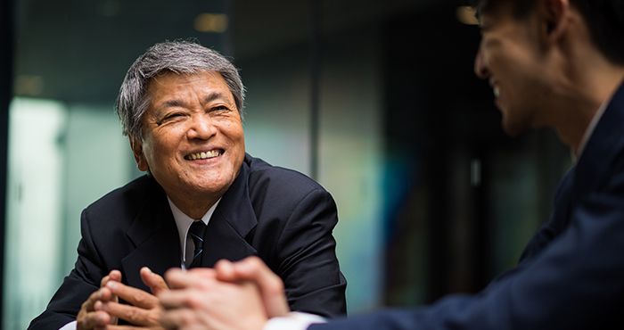 An man with gray hair and a suit smiles warmly while engaging in a conversation with a younger person in a suit. They appear to be in a professional setting, possibly an office environment.