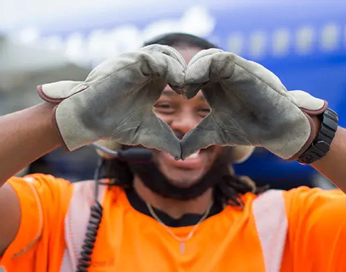 A Southwest Ramp Agent smiles while extending his hands toward the camera. His hands are in work gloves, and he's making a heart shape. Out of focus, his smile is visible through his hands. 