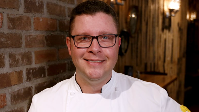 Chef Dustin smiles for the camera in their chef's coat. 