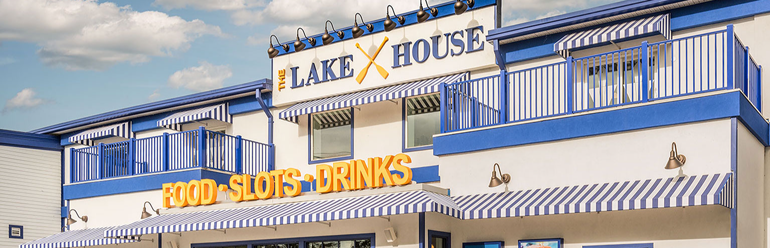 Exterior shot of The Lake House entrance, a blue and white building with three yellow signs that read, "Food," "Slots," and "Drinks." 