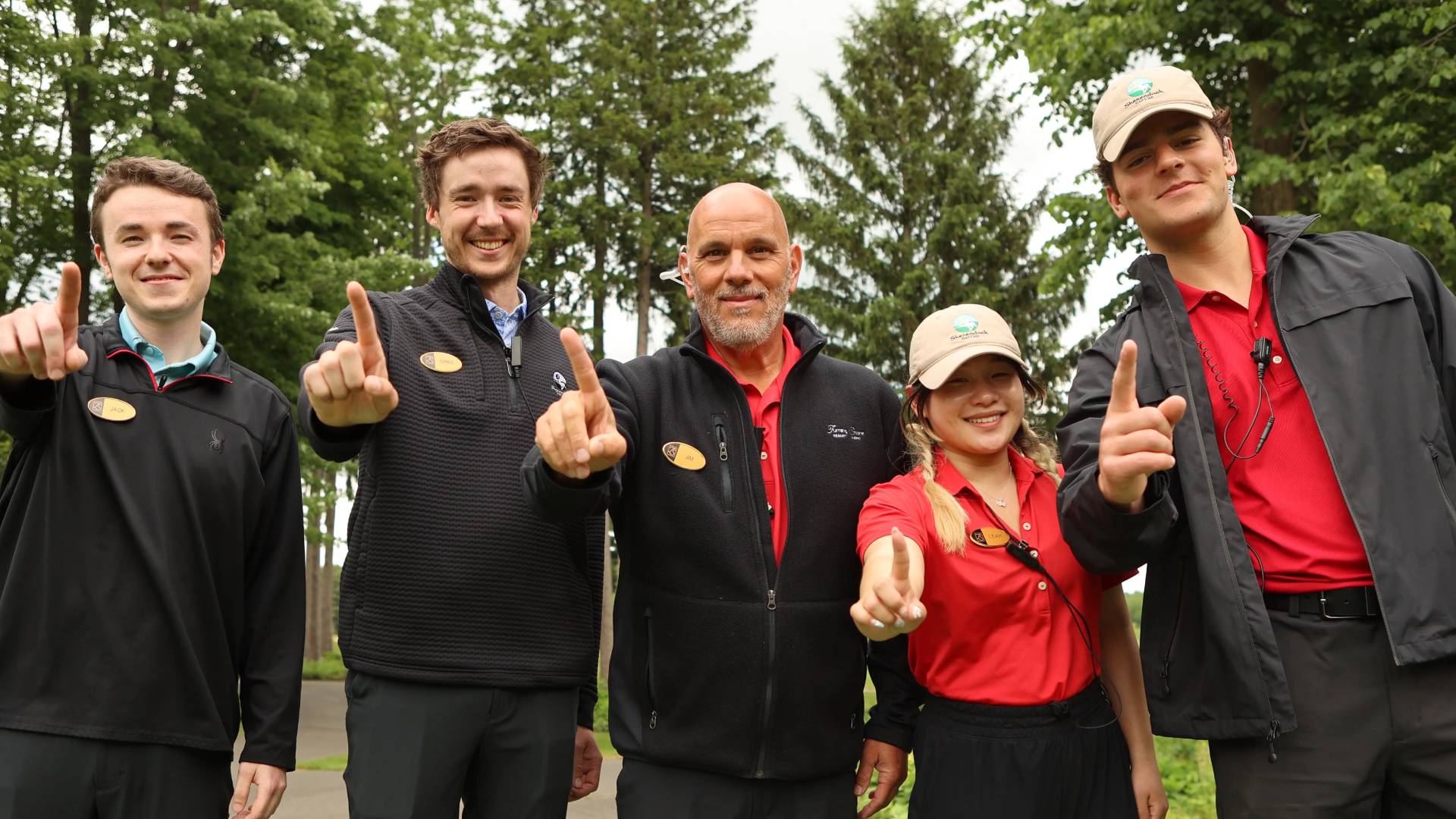 Five team members holding up their pointer fingers to show they're number one while standing outside with trees in the background. 