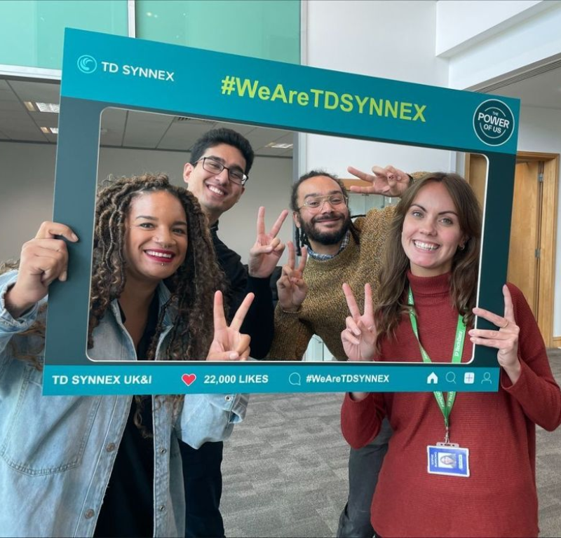 4 TD SYNNEX co-workers making peace signs with their hands and holding up a sign that says "#WeAreTDSYNNEX"