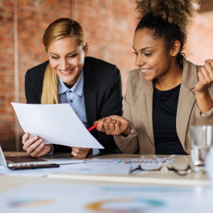 Dos compañeras de trabajo sentadas en una mesa apuntando a un trozo de papel que uno de los compañeros sostiene.