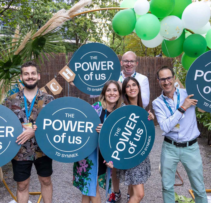 5 TD SYNNEX co-workers in an outdoors area holding up signs that read "The Power of Us"