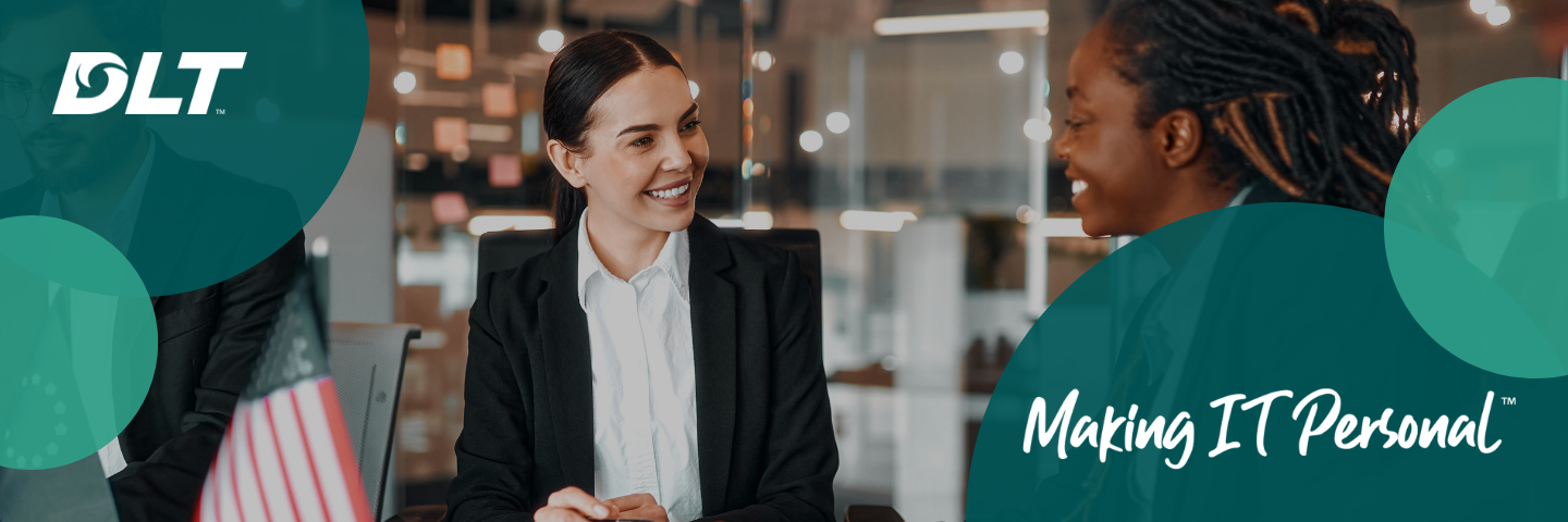 Two professional women sitting across from each other in a modern office setting, smiling and engaged in conversation, with glass walls and warm lighting in the background.