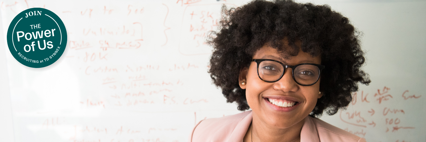 Woman smiling in front of white board with formulas written on it. Badge that reads "Join the Power of Us - Recruiting at TD SYNNEX."