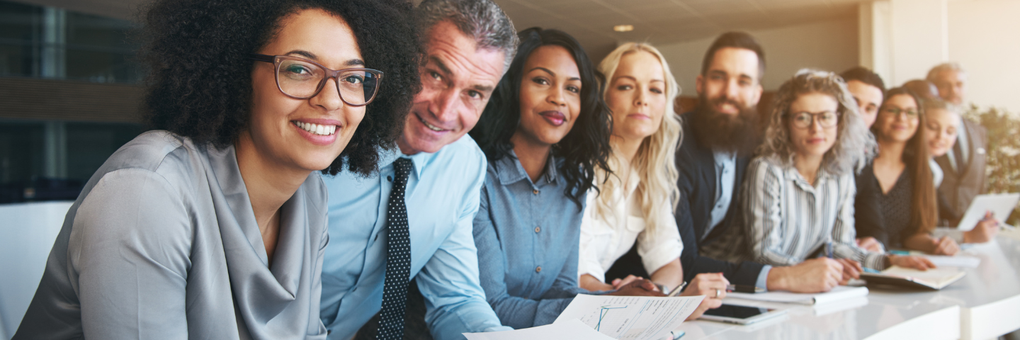 11 co-workers in professional attire smiling in an office setting with notebooks and papers in front of them