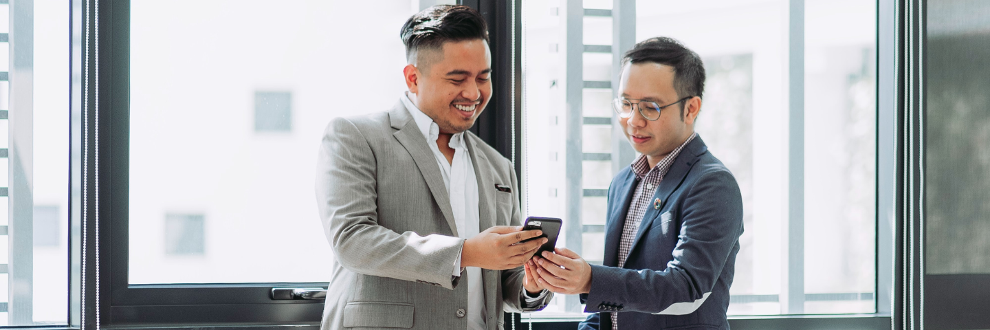 Two men in business attire holding a phone in their hands and smiling.