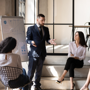 Estudiantes sentados en un entorno de oficina, mientras un orador está de pie y hace una presentación con un gran bloc de notas detrás de él.