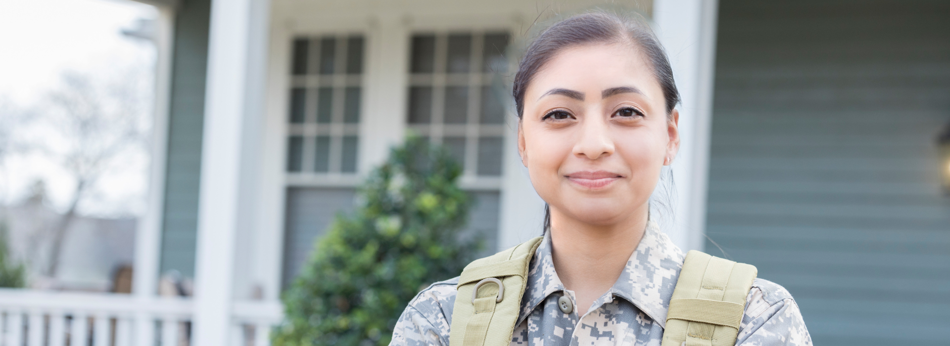 Female veteran wearing camo smiling and standing outside the front of her home.