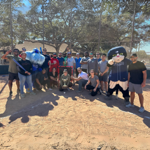 TD SYNNEX team members and 2 Tampa Bay Rays mascots posing on a wiffle ball field while holding up a trophy.