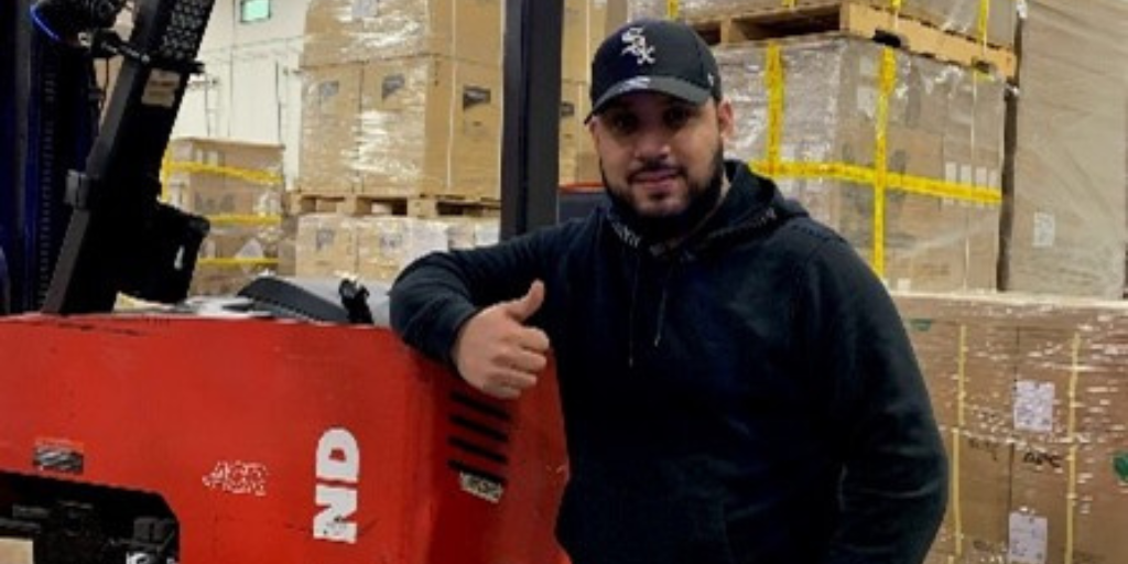 Erick giving a thumbs up while standing in our warehouse next to a forklift