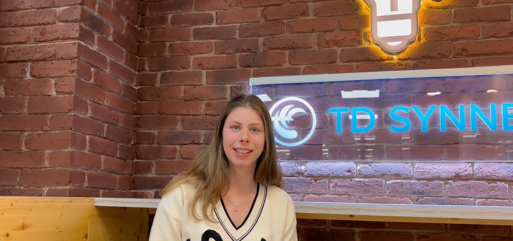 Valeria sitting in a TD SYNNEX office with a brick background and TD SYNNEX logo on display