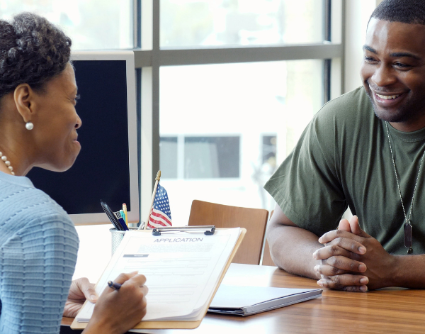 Veteran smiling while chatting with woman across from him at a table. The woman is holding a clipboard that says "Application" on it.