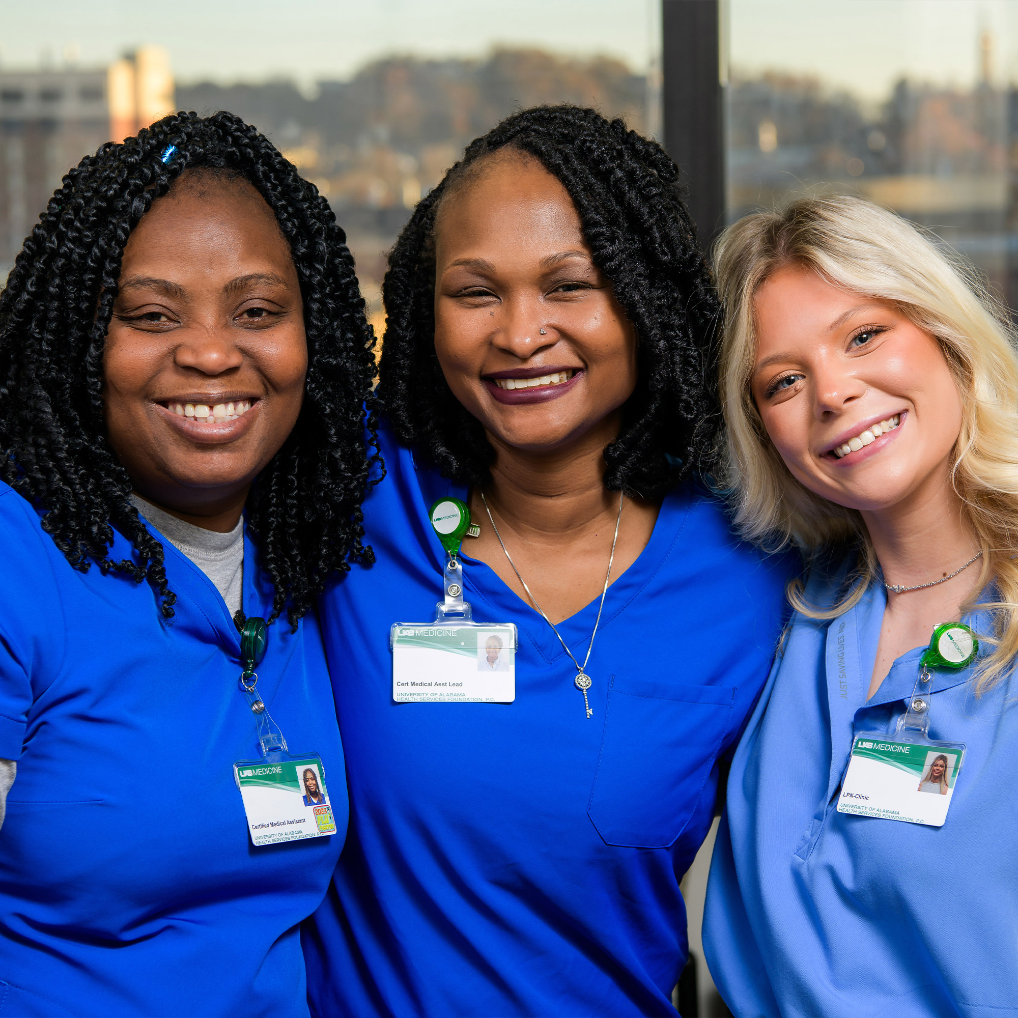 Three smiling healthcare workers in blue scrubs stand close together indoors, wearing name badges and posing in front of a window with a blurred cityscape in the background.
