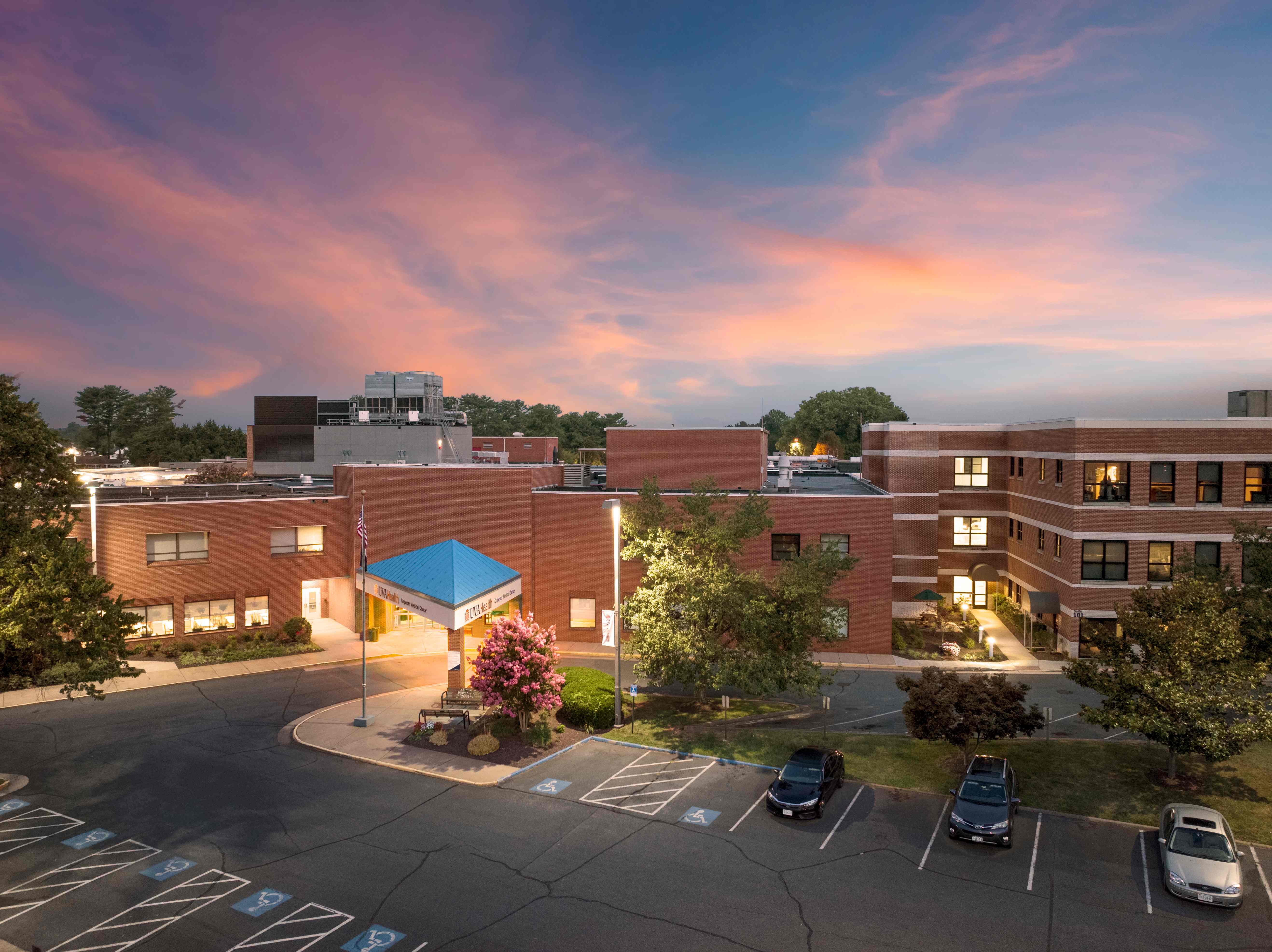 UVA Health Culpeper Medical Center at sunset, with a pink and purple sky, blue entrance canopy, brick exterior, and parking in front.