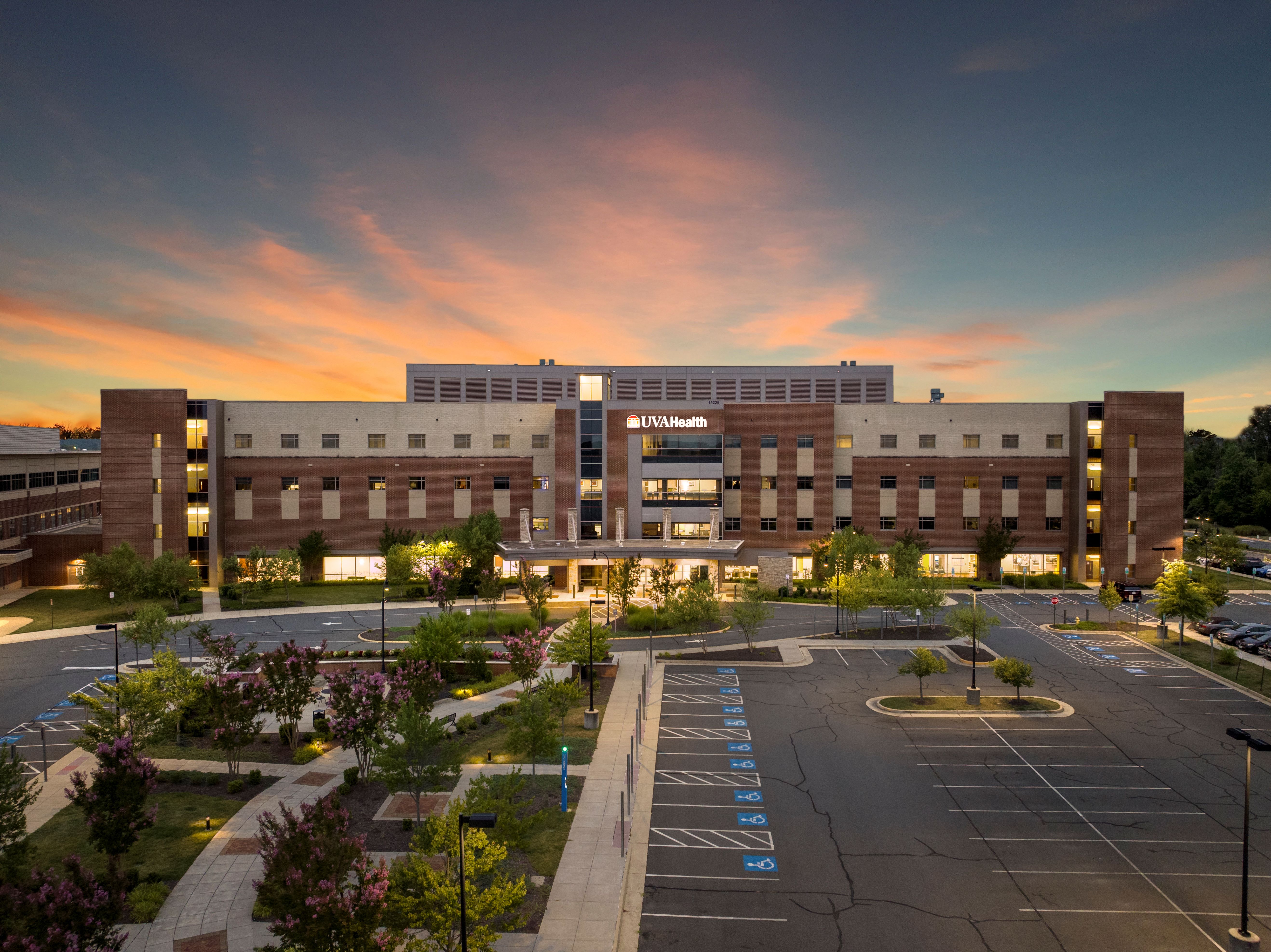 UVA Health Haymarket Medical Center at sunset, with an orange and pink sky, illuminated entrance, landscaped walkways, and parking in front.