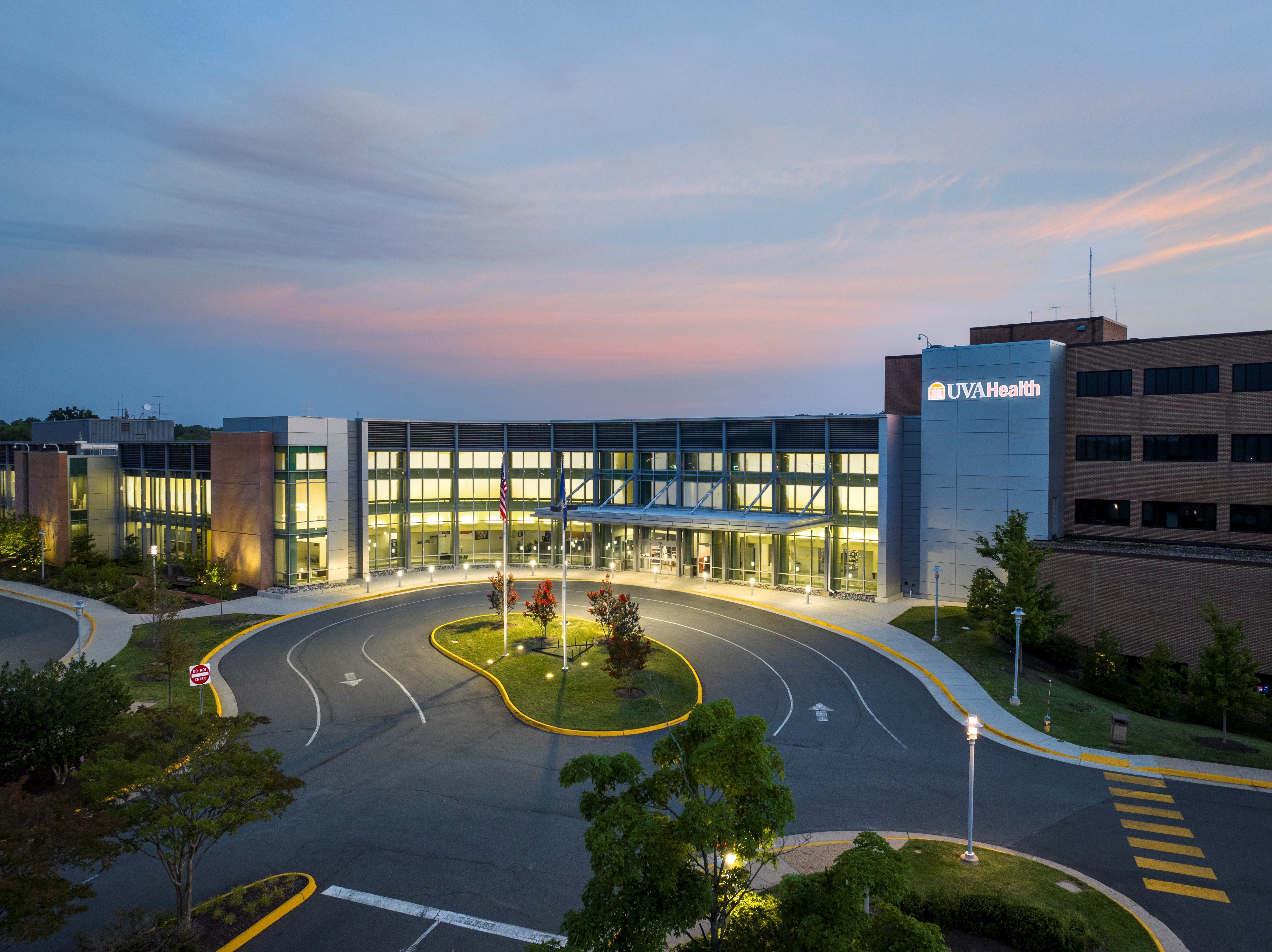 UVA Health Prince William Medical Center at sunset, with an illuminated glass entrance and a circular driveway surrounding flagpoles and landscaping.