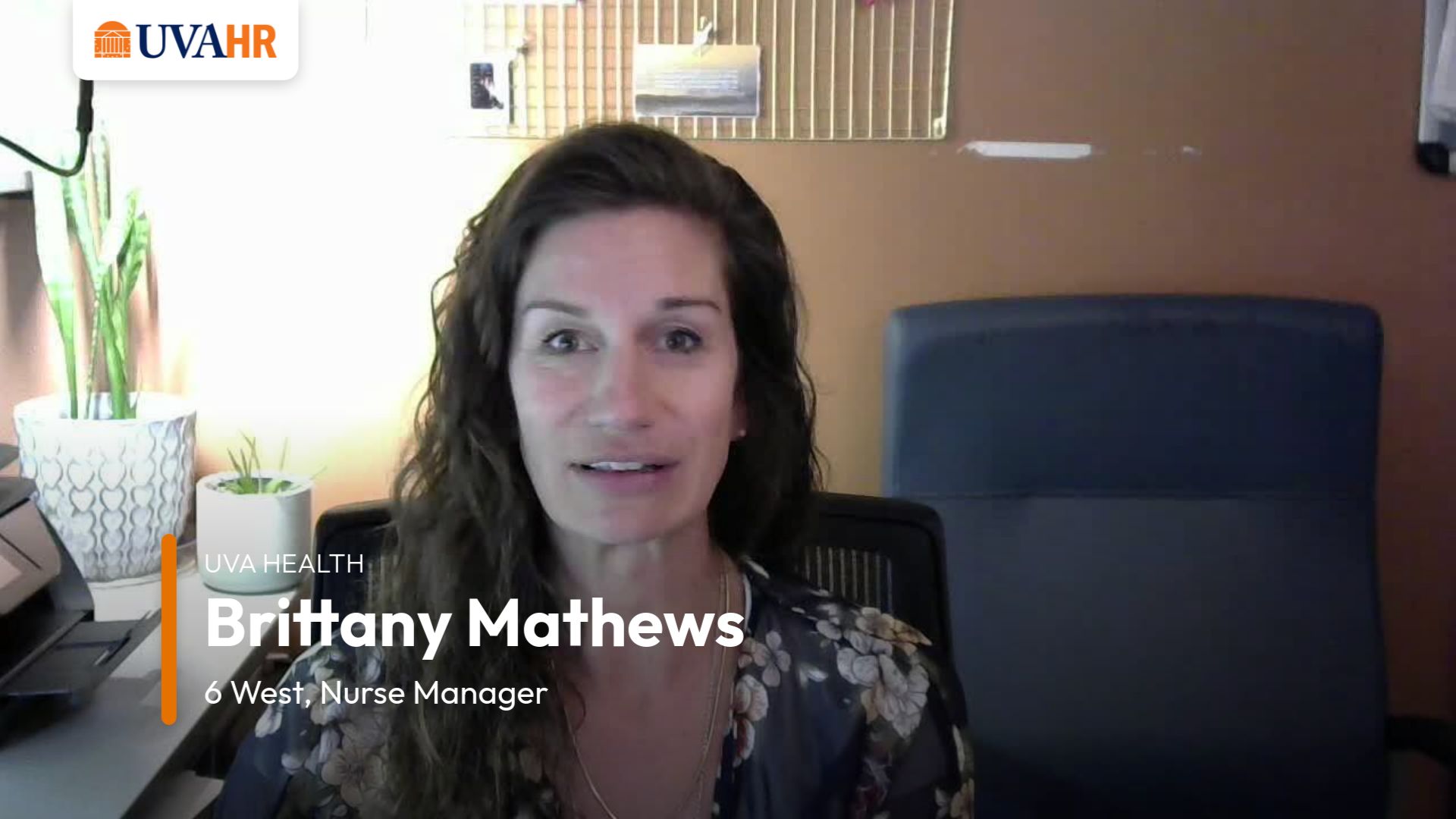 Person speaks directly to the camera in room, table in the background has two plants and there is a blue chair in the background on the right.