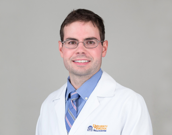 A person wearing a white University of Virginia Health System lab coat, a blue collared shirt, and a striped tie, posed against a plain light gray background.