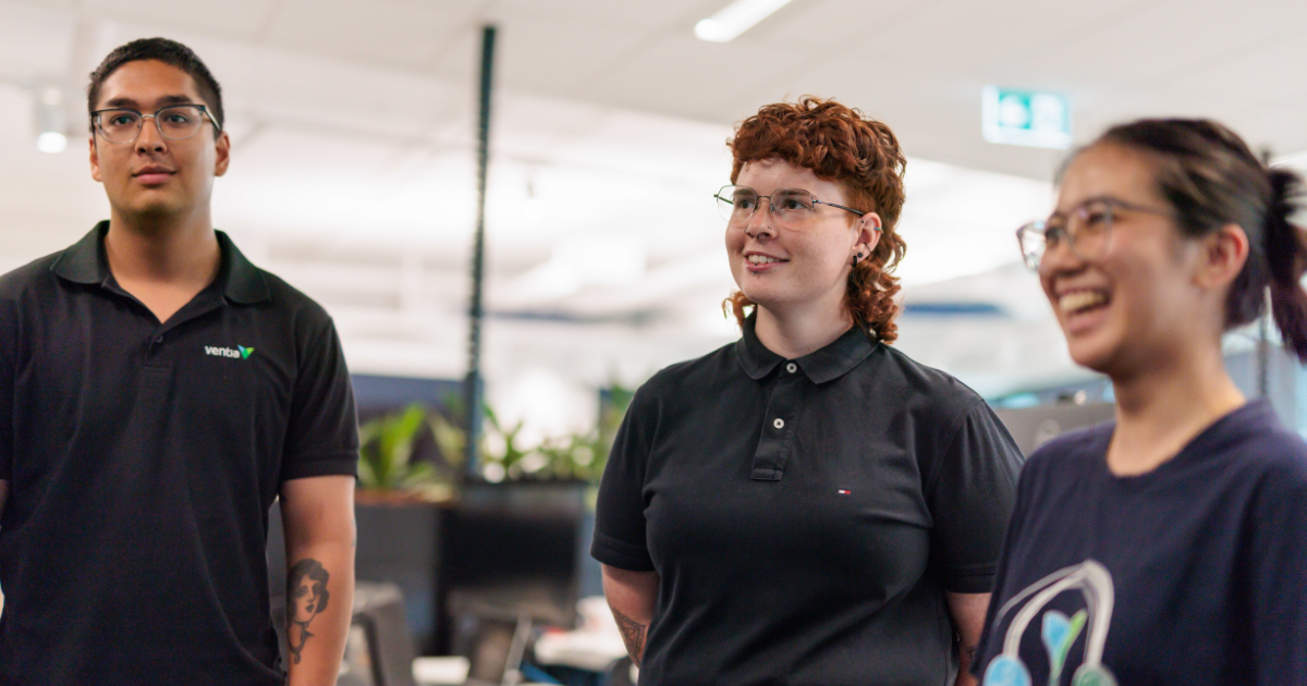 : Three people standing in an office environment with blurred faces. The person on the left is wearing a black polo shirt with a logo, the person in the middle has curly hair and is wearing a black polo shirt, and the person on the right is wearing a dark blue t-shirt with a design. The background shows office desks and plants