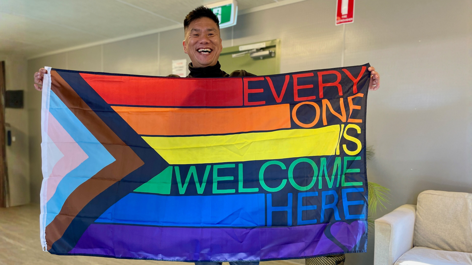 man smiling holding a rainbow flag with the text everyone is welcome here