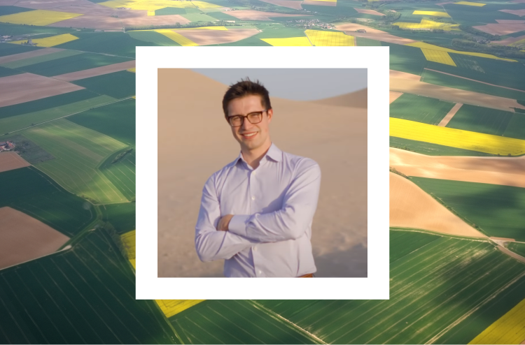 A smiling man with short dark hair and glasses stands with his arms crossed, wearing a light blue dress shirt. His photo is framed within a white border, set against an aerial view of patchwork farmland with various shades of green and yellow fields, creating a scenic agricultural landscape. The background contrasts with the sandy desert visible in the photo within the white border.