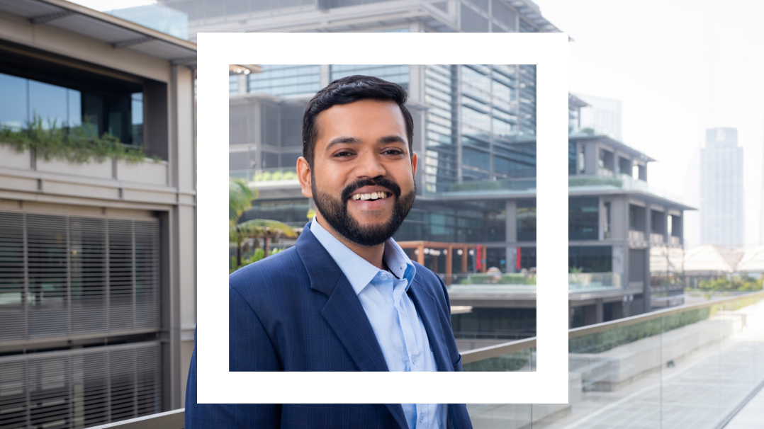 A man with a beard, wearing a blue suit and light blue shirt, smiles while standing outdoors in a modern urban environment with tall buildings and glass windows in the background. The image is framed within a white rectangular overlay.