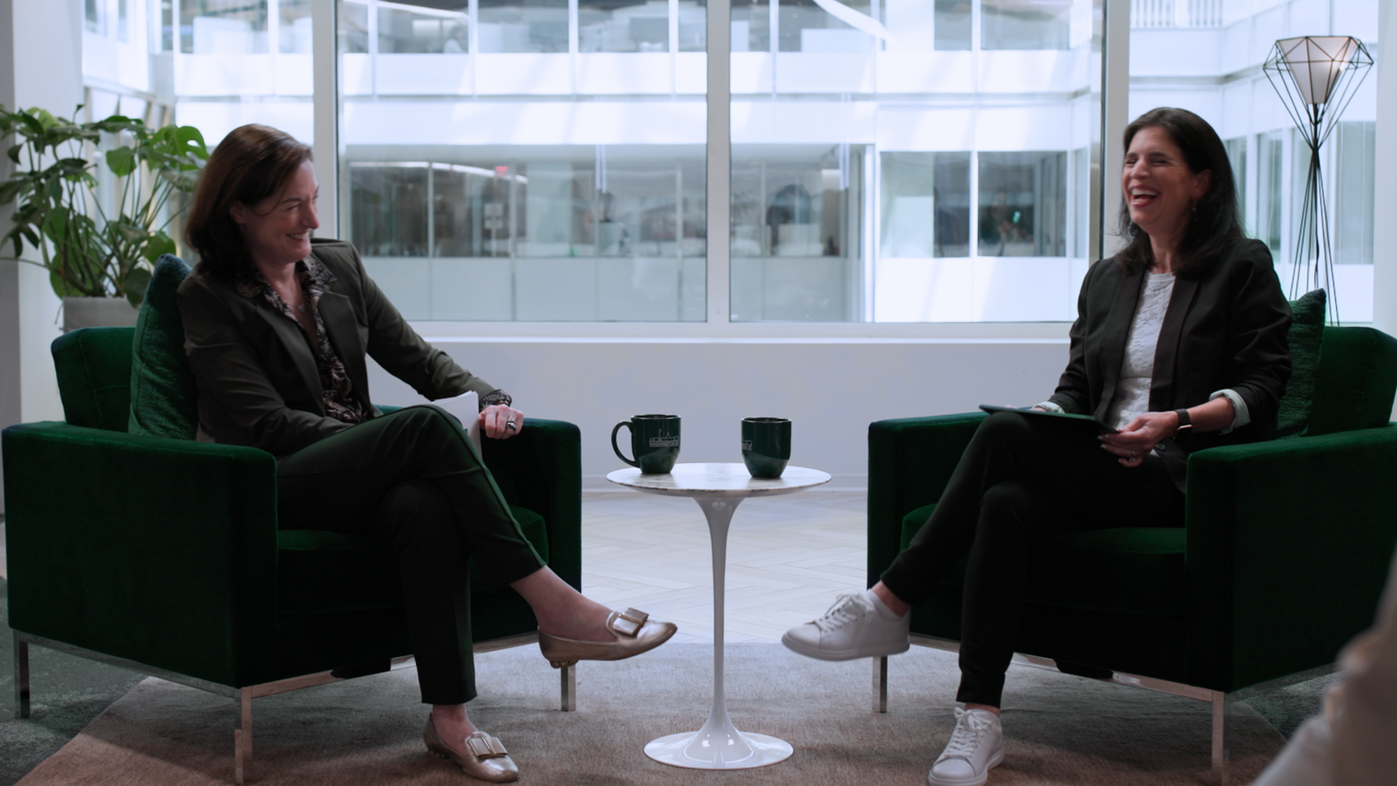 The thumbnail photo shows two women seated in green armchairs facing each other in a brightly lit, modern indoor space with large windows in the background. They are engaged in a conversation, both smiling. A small round white table is positioned between them with a pair of headphones placed on it. The woman on the left is dressed in dark clothing with light-colored shoes, and the woman on the right is wearing dark pants and a jacket with white sneakers. A white play button icon is centered on the image, indicating that it is a video thumbnail.